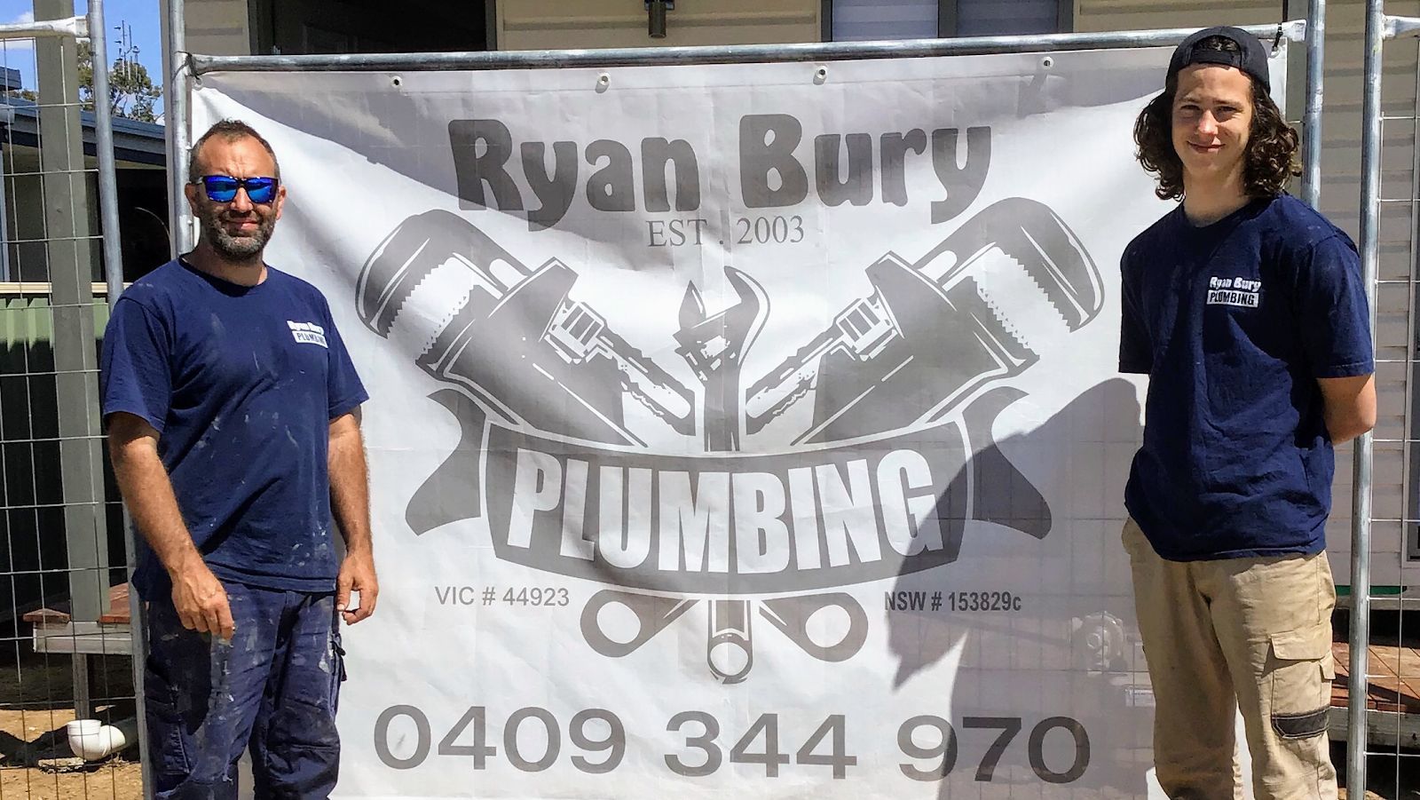 Two of the Ryan Bury Plumbing crew in front of the company banner on a fenced job site